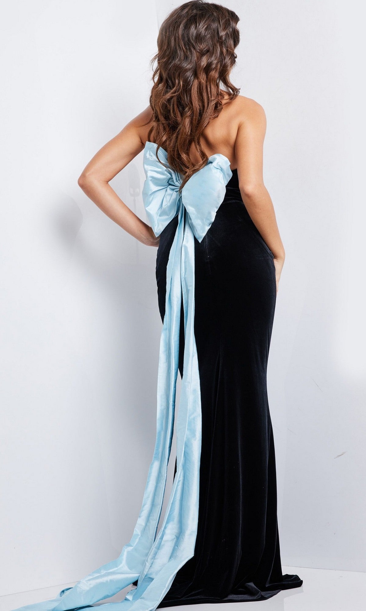 A woman faces away from the camera in the Jovani 26227 long prom dress, showcasing its strapless velvet design with a signature oversized back bow and a flowing light blue sash.